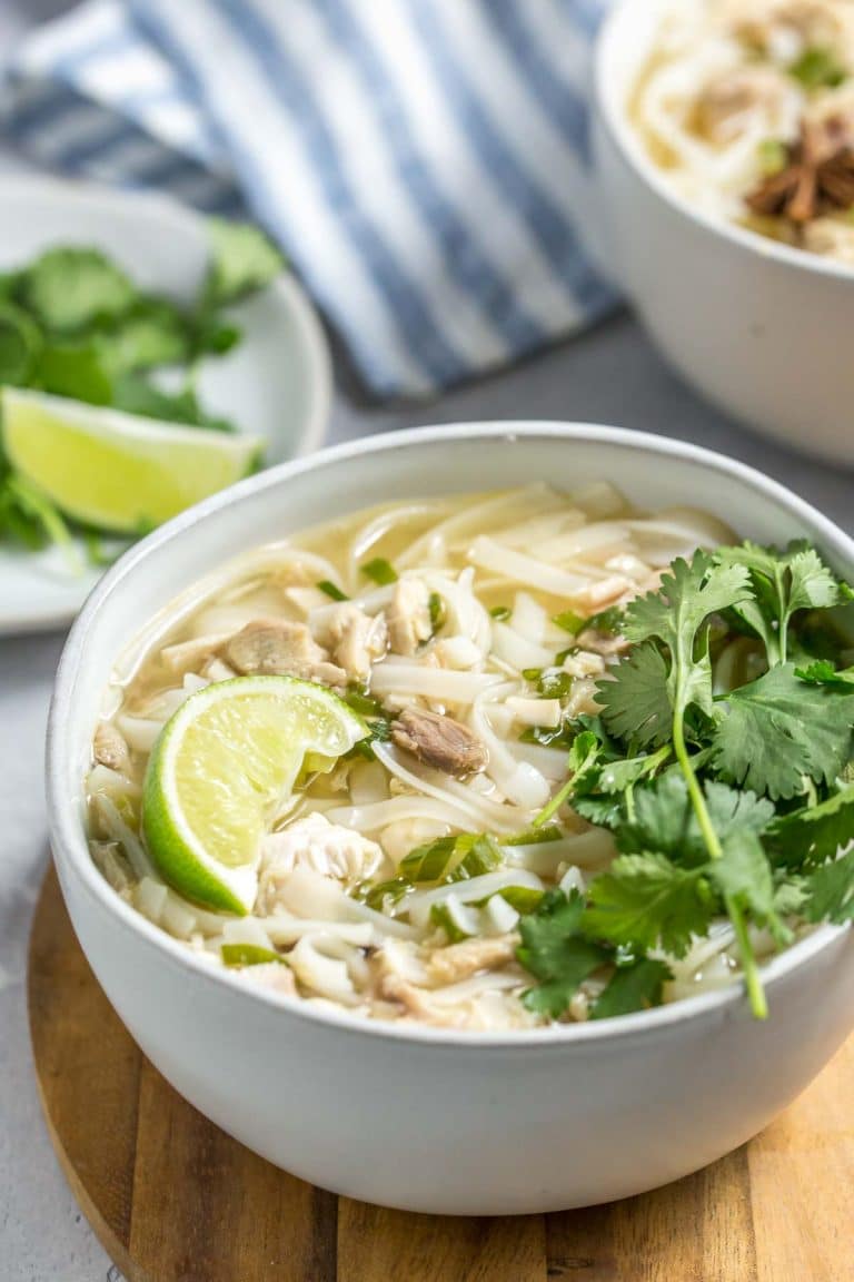 closeup shot of leftover turkey pho in a bowl with chopsticks next to it, topped with fresh herbs, a lime wedge and star anise, extra bowls of soup and herbs in the background