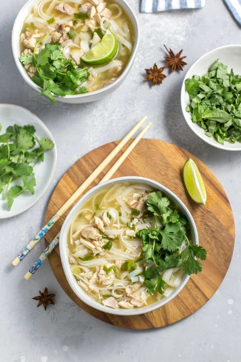 Overhead shot of leftover turkey pho in a bowl with chopsticks next to it, topped with fresh herbs, a lime wedge and star anise, extra bowls of soup and herbs in the background