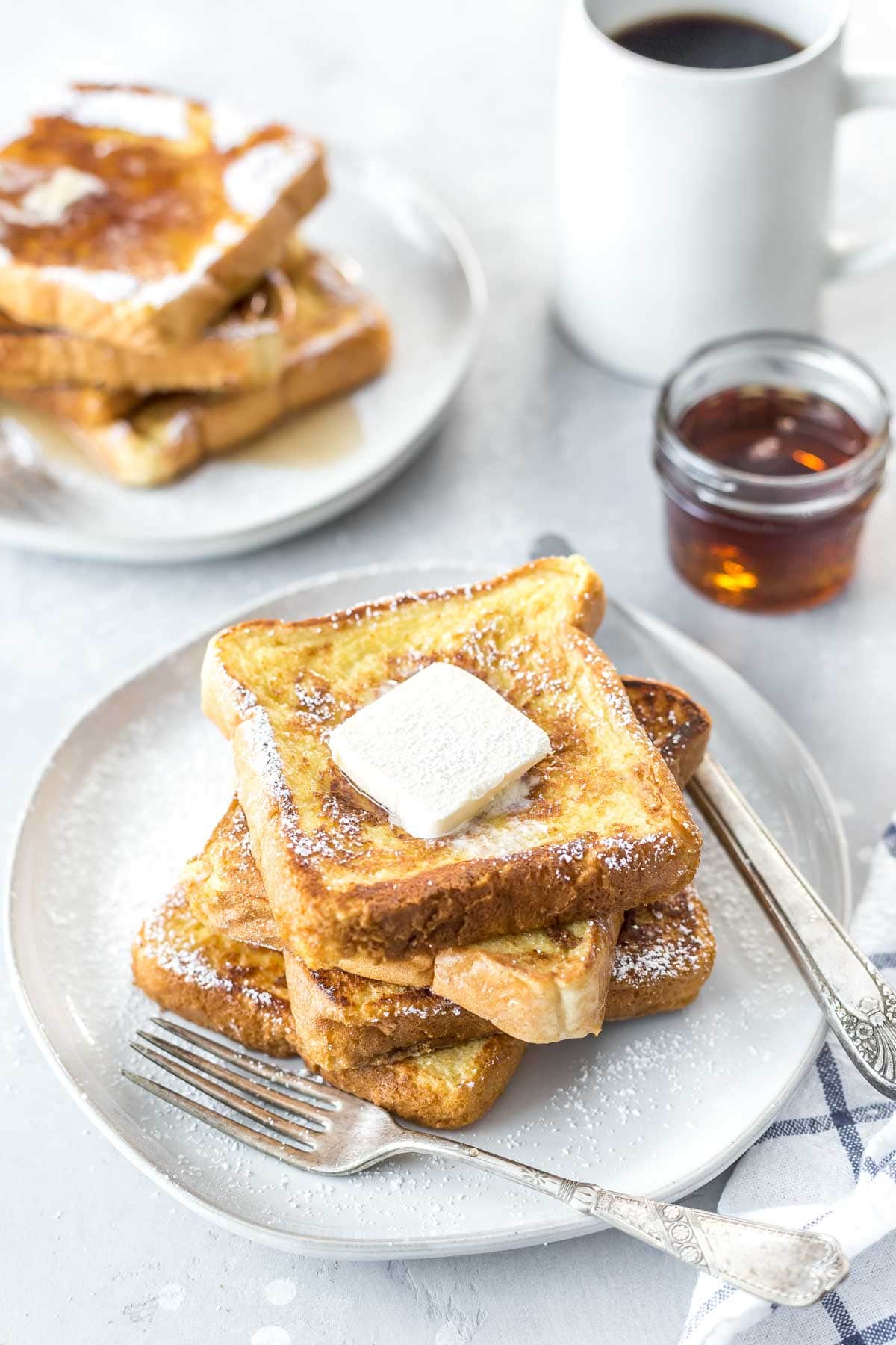 dairy free french toast topped with syrup, powdered sugar and vegan butter, a second stack of no milk french toast in the background with coffee and a small mason jar of syrup