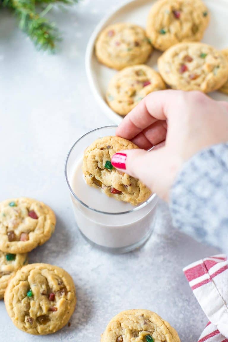 hand dipping a fruitcake cookies into a glass of cashew milk with more cookies scattered around the glass and a red and ivory striped napkin and pine leaves in the background