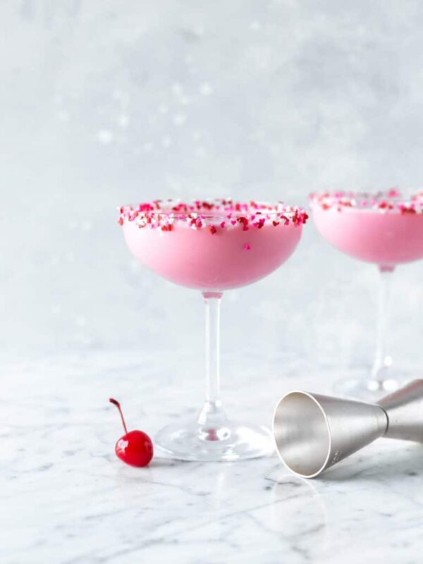 two pink squirrel cocktails in coupe glasses rimmed with miniature heart sprinkles, a single maraschino cherry at the base of the first glass and a cocktail jigger on the other side