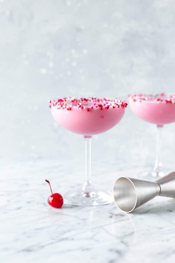 two pink squirrel cocktails in coupe glasses rimmed with miniature heart sprinkles, a single maraschino cherry at the base of the first glass and a cocktail jigger on the other side