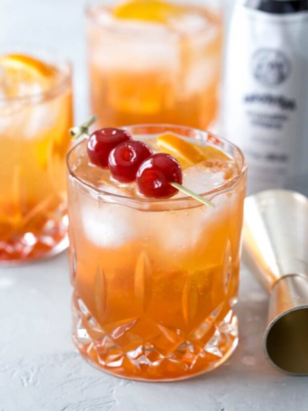 three wisconsin brandy old fashioned cocktails, the front cocktail topped with a skewer of three maraschino cherries, a jigger and bottle of bitters beside the glasses