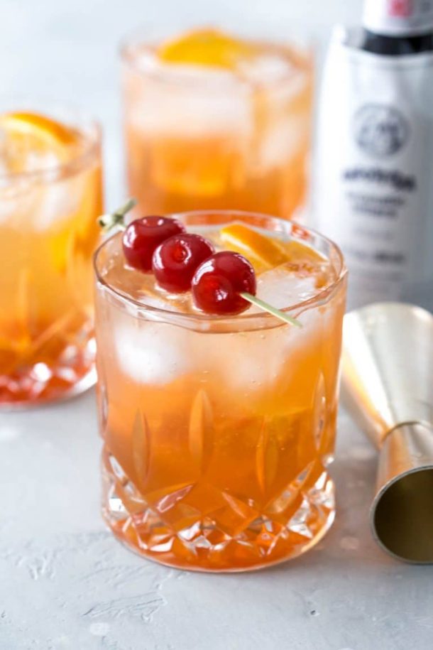 three wisconsin brandy old fashioned cocktails, the front cocktail topped with a skewer of three maraschino cherries, a jigger and bottle of bitters beside the glasses
