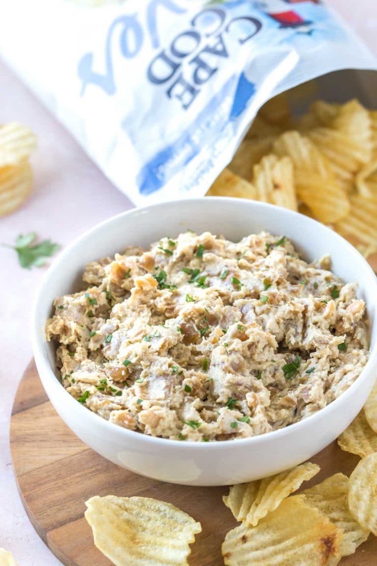 caramelized onion dip in a white bowl on a wooden serving board with a bag of cape cod waves chips in the background, chips surrounding the bowl