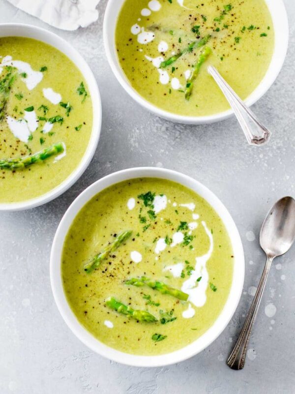 three bowls of creamy asparagus soup in a white bowl, garnished with asparagus tips, parsley and drizzles of coconut milk