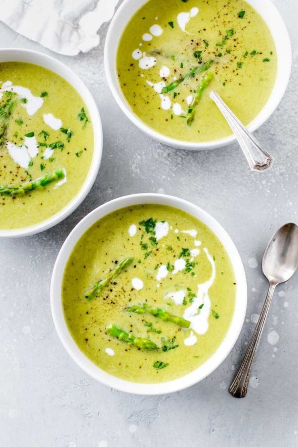 three bowls of creamy asparagus soup in a white bowl, garnished with asparagus tips, parsley and drizzles of coconut milk