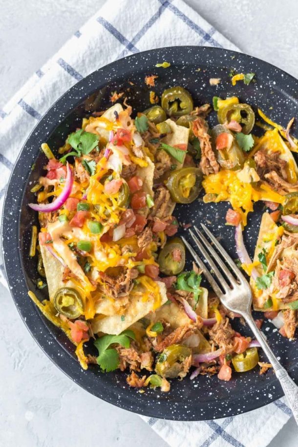 half eaten BBQ pulled pork nachos in a black speckled pie plate on a white and blue dish towel with lime wedges to the side and a small bowl of cilantro