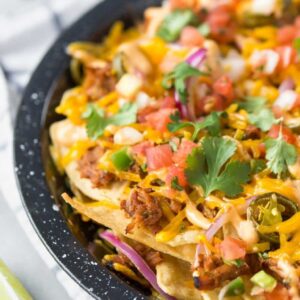 closeup of BBQ pulled pork nachos in a black speckled pie plate on a white and blue dish towel with lime wedges to the side and a small bowl of cilantro