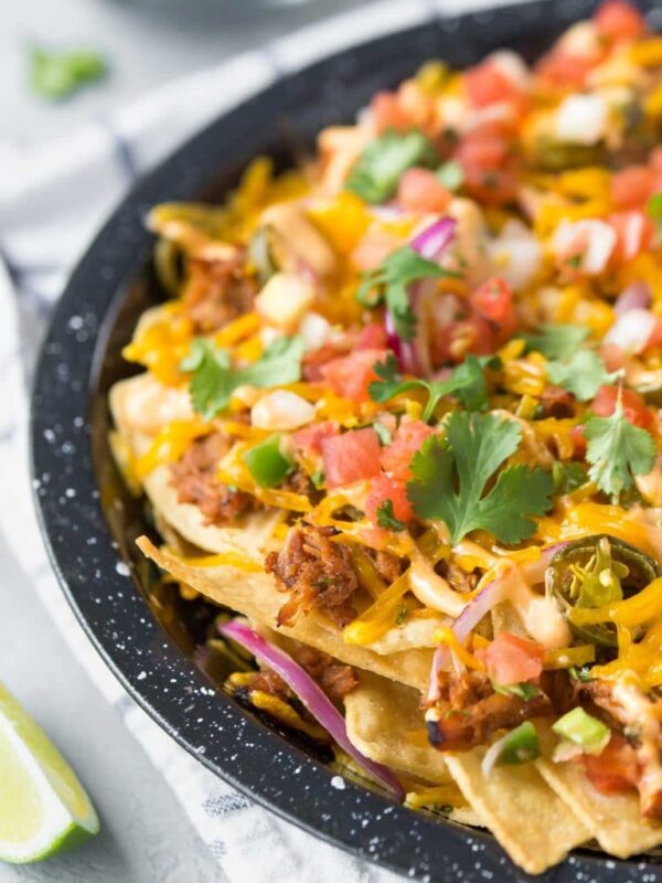 closeup of BBQ pulled pork nachos in a black speckled pie plate on a white and blue dish towel with lime wedges to the side and a small bowl of cilantro