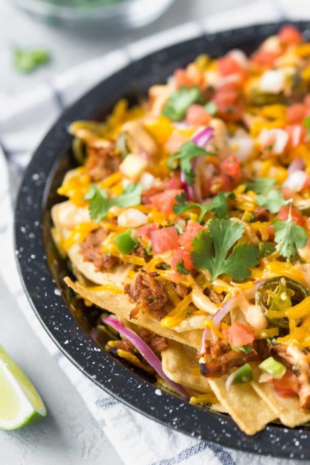 closeup of BBQ pulled pork nachos in a black speckled pie plate on a white and blue dish towel with lime wedges to the side and a small bowl of cilantro