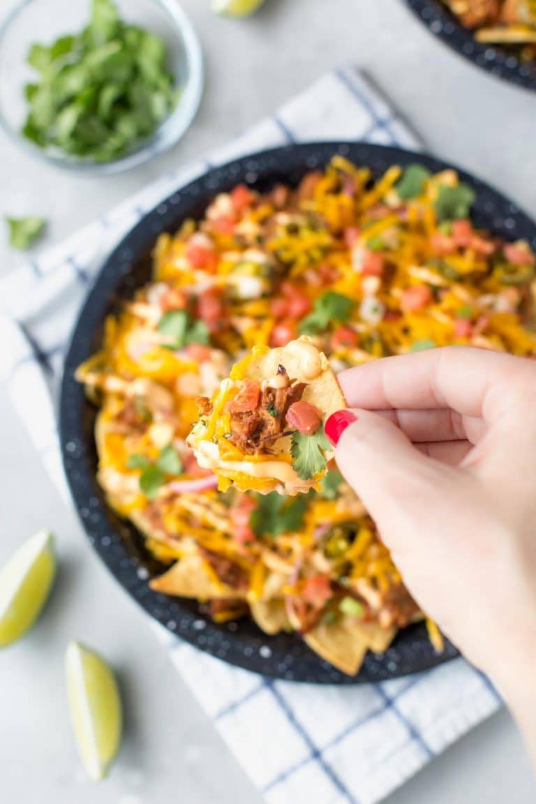 hand holding a tortilla chip topped BBQ pulled pork nachos above a plate of nachos in a black speckled pie plate on a white and blue dish towel with lime wedges to the side and a small bowl of cilantro