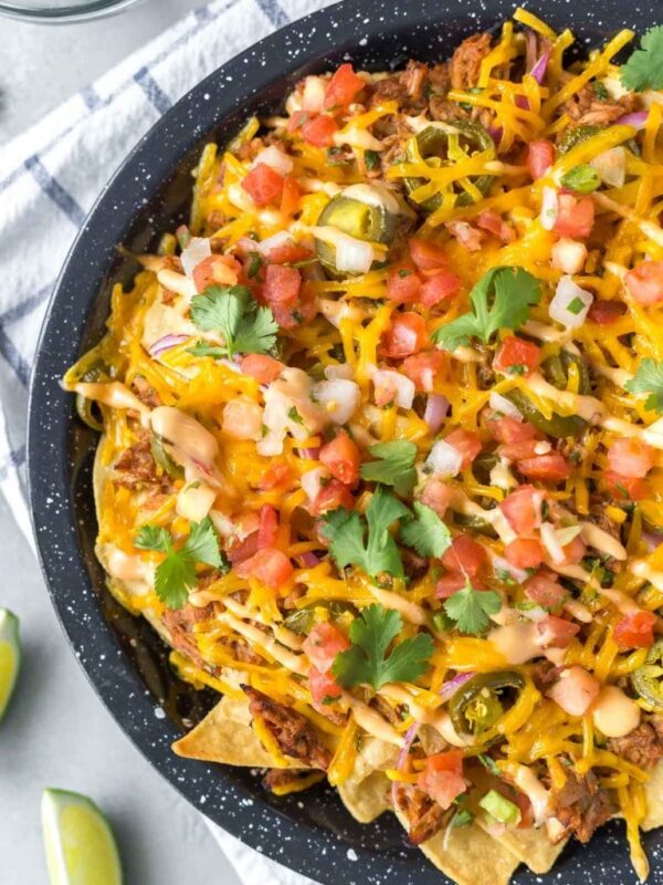 closeup of BBQ pulled pork nachos in a black speckled pie plate on a white and blue dish towel with lime wedges to the side and a small bowl of cilantro