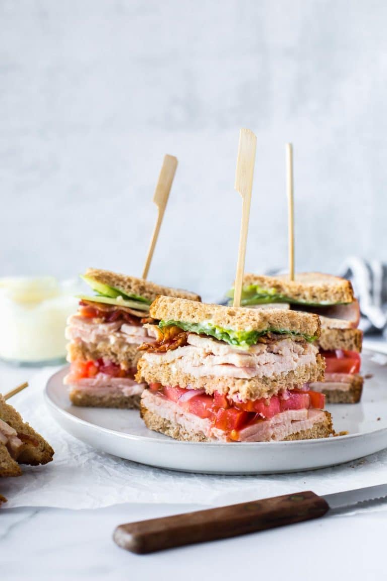 three sections of a turkey club sandwich on a gray plate with wooden skewers in the top, a jar of aioli and a black and white striped cloth napkin in the background and a fourth section of the club sandwich on its side in the foreground
