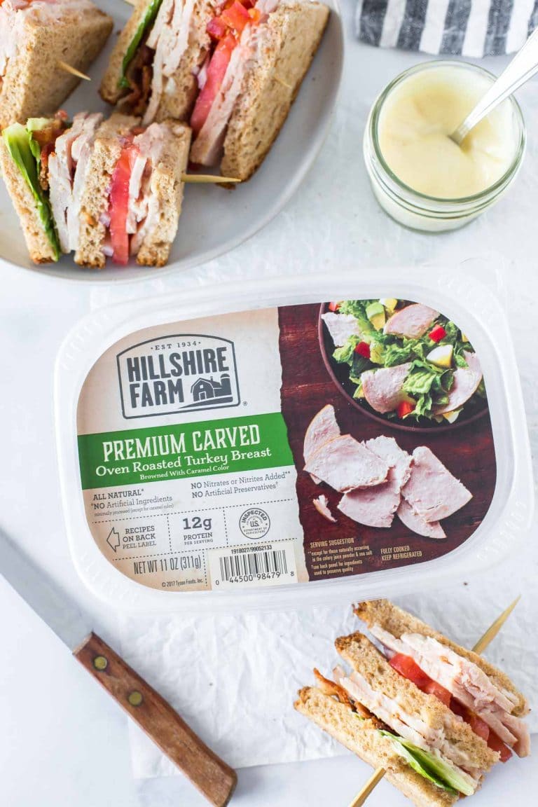 a package of turkey lunch meat, surrounded by a jar of aioli, a bread knife, and cut sections of a turkey club sandwich on wooden skewers