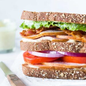 closeup of a turkey club sandwich on crinkled parchment paper with a bread knife laying besides it, a small jar of aioli in the background