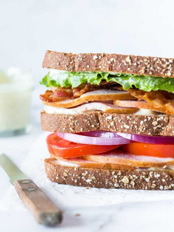 closeup of a turkey club sandwich on crinkled parchment paper with a bread knife laying besides it, a small jar of aioli in the background