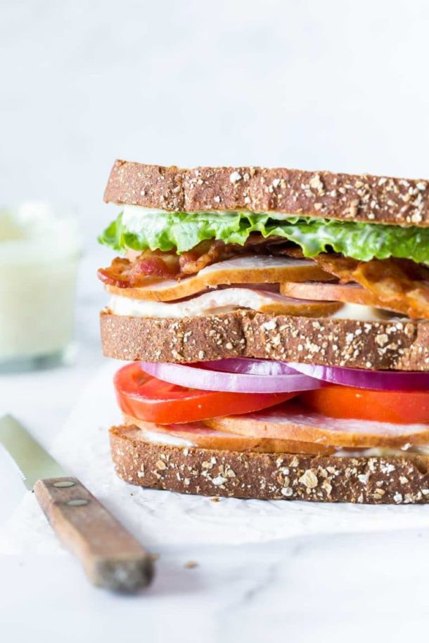 closeup of a turkey club sandwich on crinkled parchment paper with a bread knife laying besides it, a small jar of aioli in the background