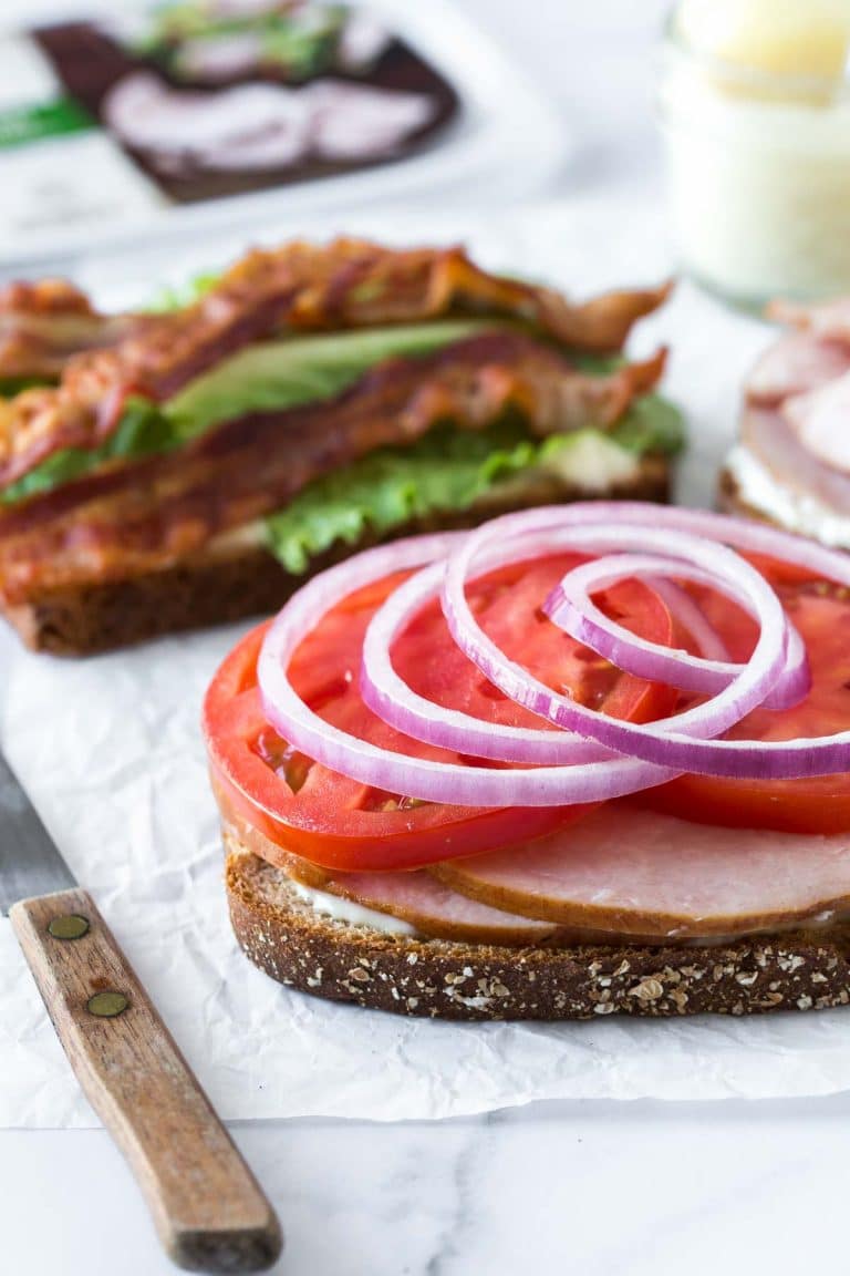how to make a club sandwich - toasted bread topped with sliced turkey, tomatoes and red onion, behind it is a second slice of toasted bread topped with aioli, lettuce and three bacon slices