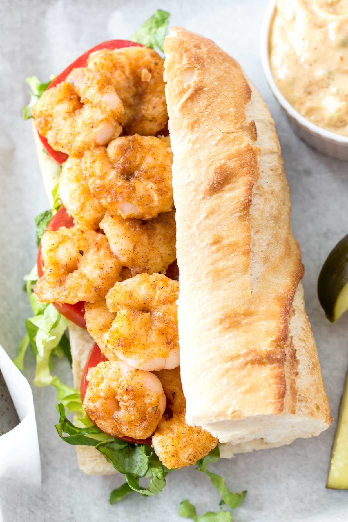 A shrimp po boy sandwich, featuring pan fried shrimp, shredded green lettuce and sliced tomatoes on a demi-baguette, homemade Cajun remoulade sauce in a white ramekin and two pickle spears to the side on a baking sheet lined with parchment paper