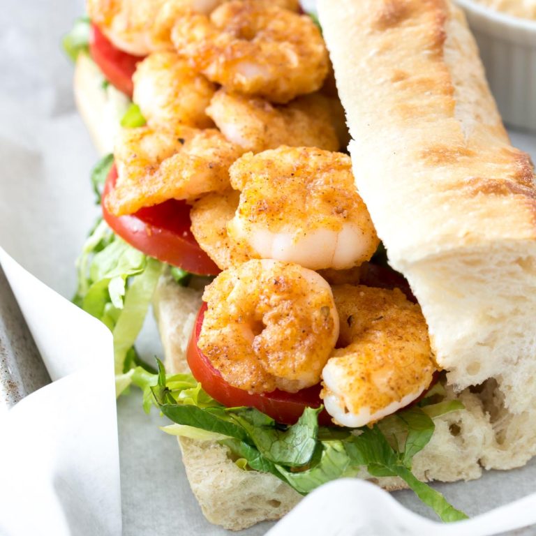 A shrimp po boy sandwich, featuring pan fried shrimp, shredded green lettuce and sliced tomatoes on a demi-baguette, homemade Cajun remoulade sauce in a white ramekin to the side on a baking sheet lined with parchment paper