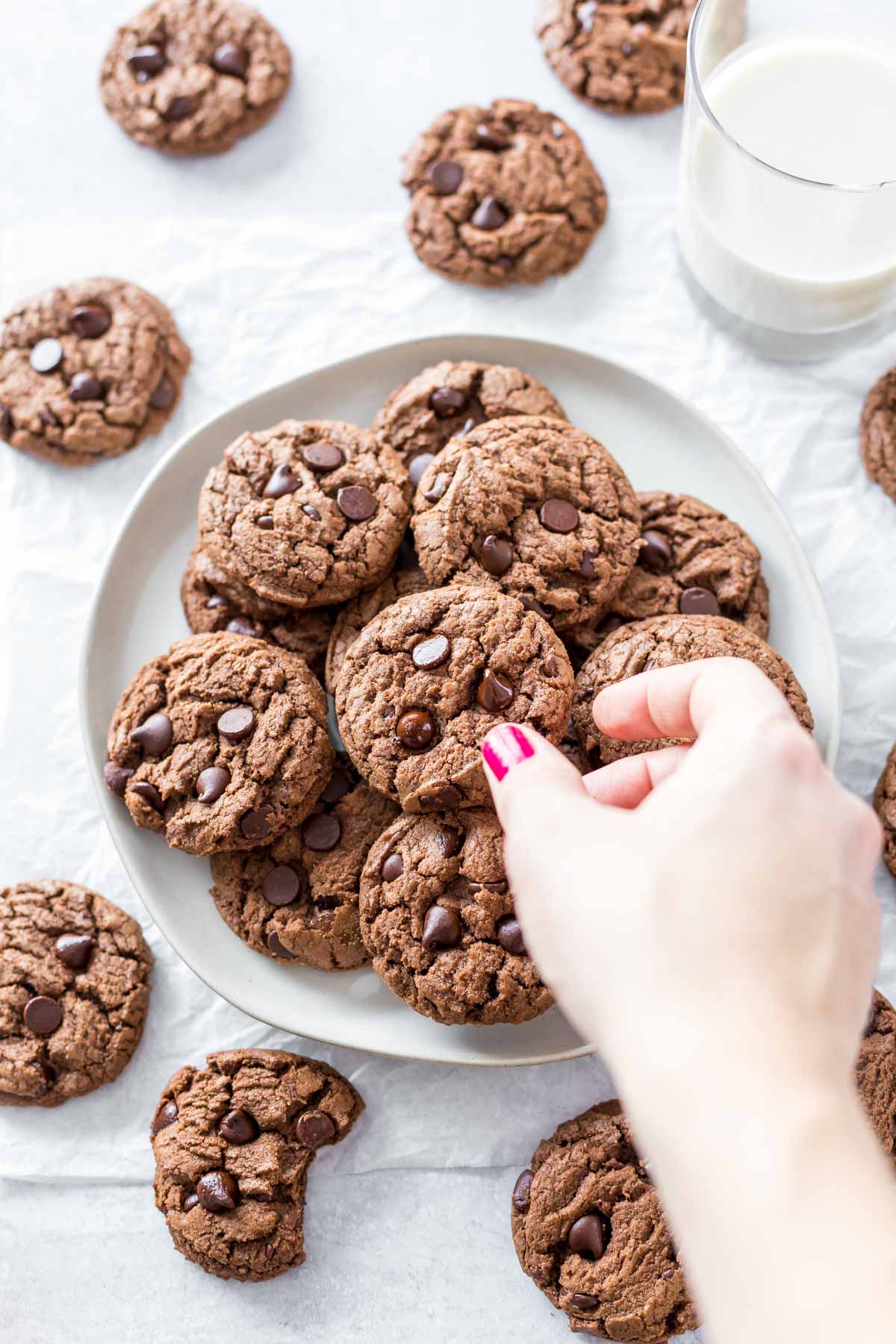 a hand reaching for chocolate chocolate chip cookies on a plate surrounded by more cookies and short glass of almond milk