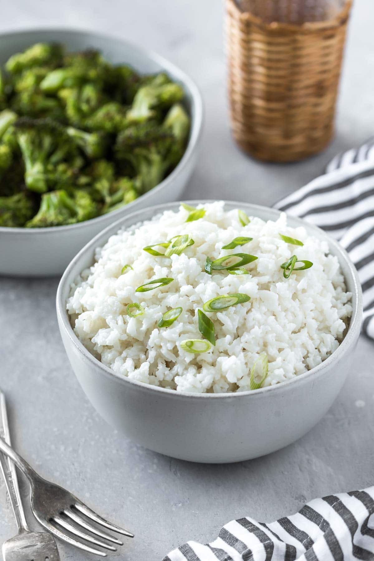 coconut rice recipe in a serving bowl topped with sliced green onions on a gray surface with forks, a striped napkin and a bowl of broccoli
