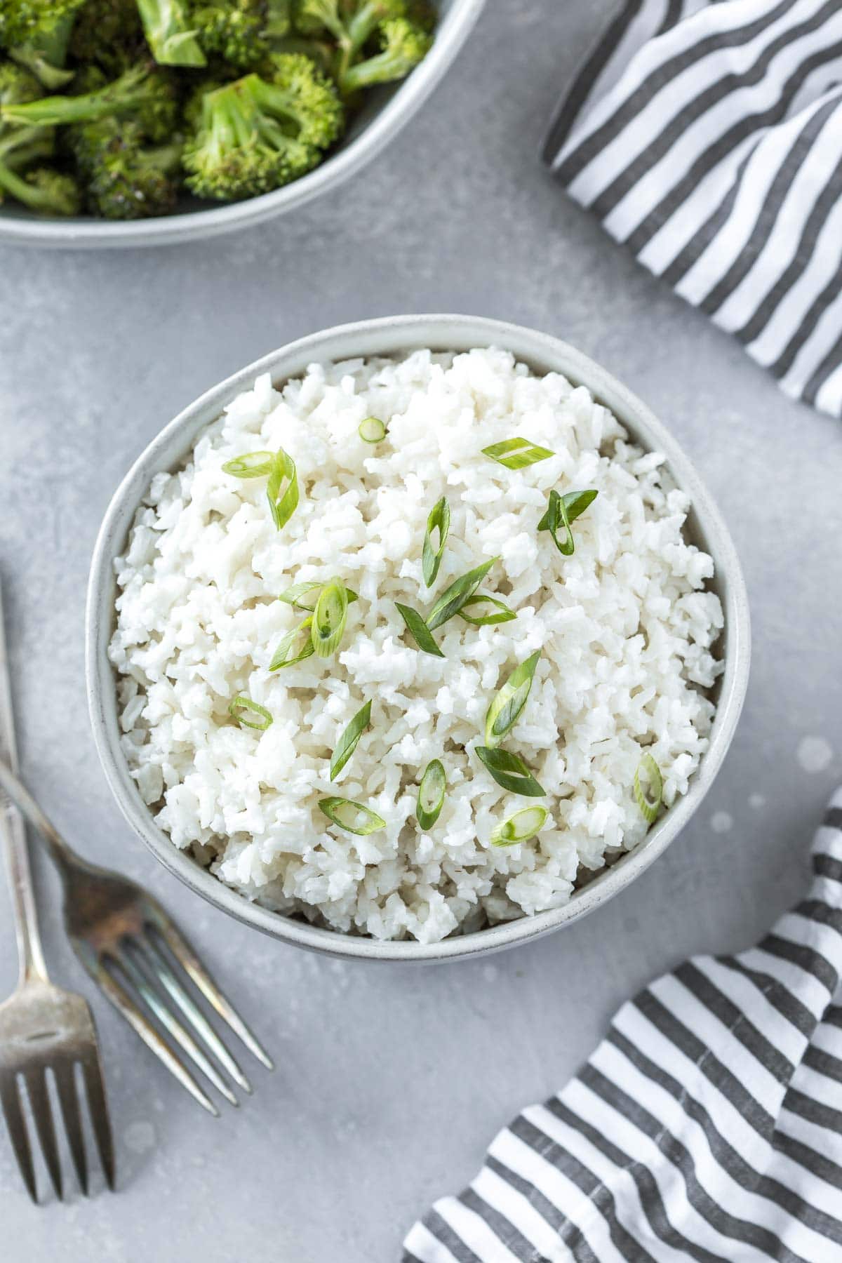 coconut rice recipe in a serving bowl topped with sliced green onions on a gray surface with forks, a striped napkin and a bowl of broccoli
