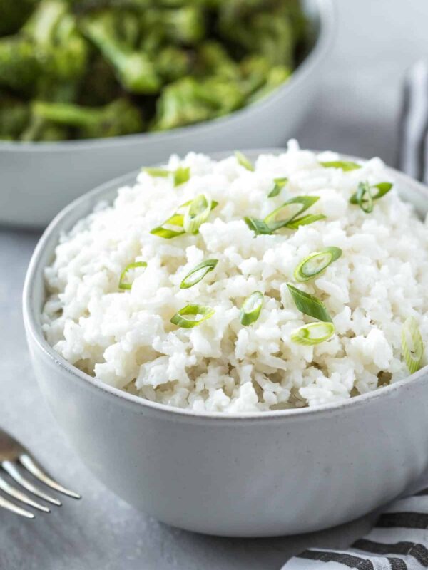easy coconut rice recipe in a serving bowl topped with sliced green onions on a gray surface with forks, a striped napkin and a bowl of broccoli