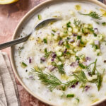 A bowl of creamy Easy Vegan Tzatziki topped with chopped cucumber, red onion, fresh dill, black pepper, and a drizzle of olive oil, with a spoon in the bowl and a lemon wedge nearby.