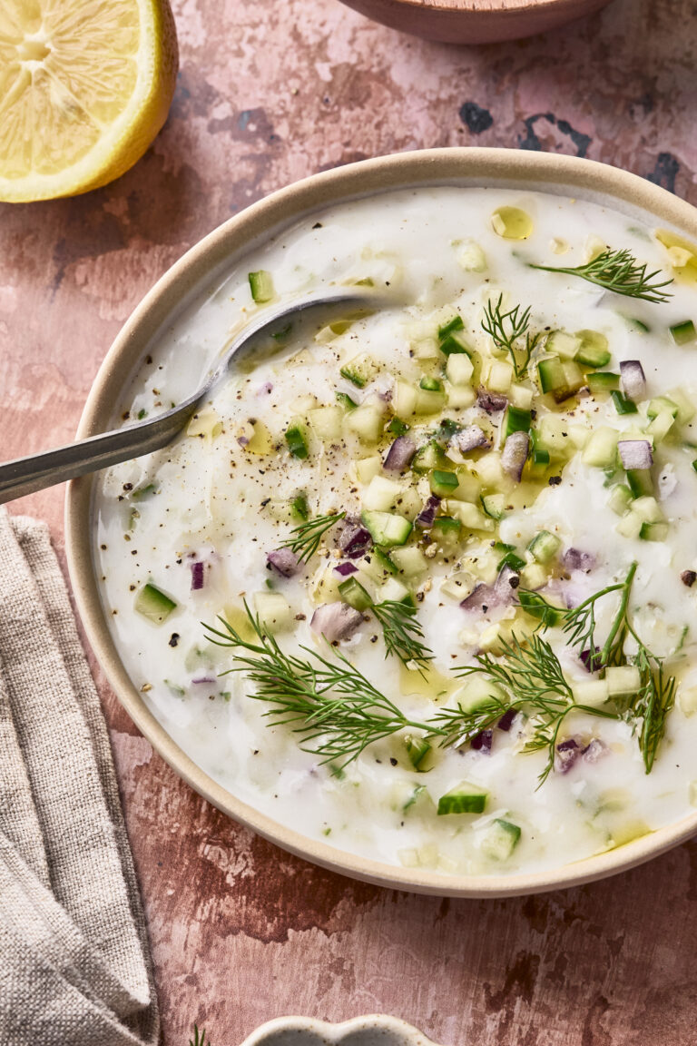 A bowl of creamy Easy Vegan Tzatziki topped with chopped cucumber, red onion, fresh dill, black pepper, and a drizzle of olive oil, with a spoon in the bowl and a lemon wedge nearby.