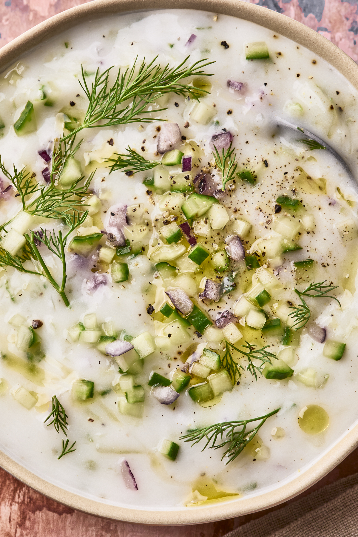 Close up photo of a bowl of creamy dairy free tzatziki sauce is garnished with chopped cucumber, red onion, fresh dill, black pepper, and a drizzle of olive oil.
