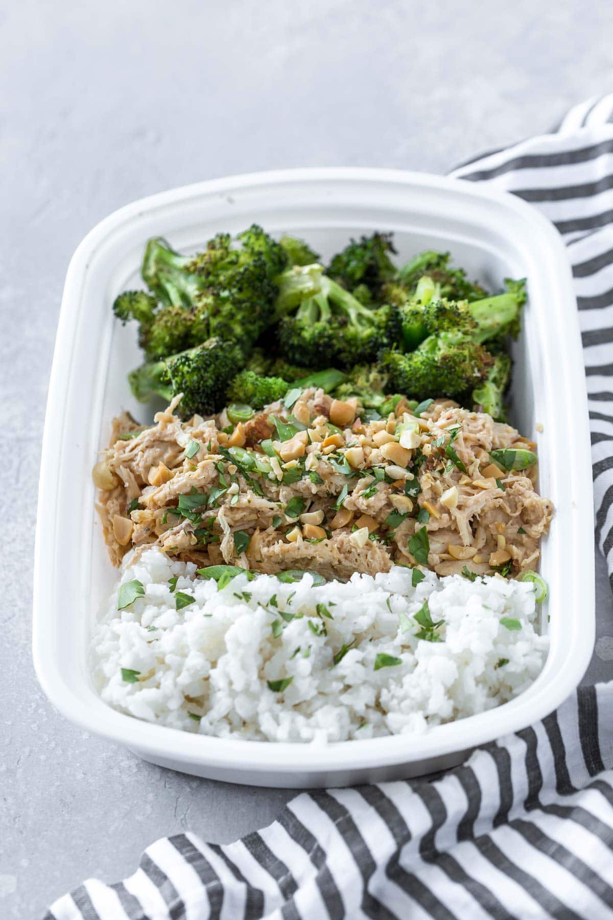 a white takeout container with coconut rice, shredded thai peanut chicken garnished with cilantro and peanuts, and roasted broccoli, on a gray background with a black and white striped napkin