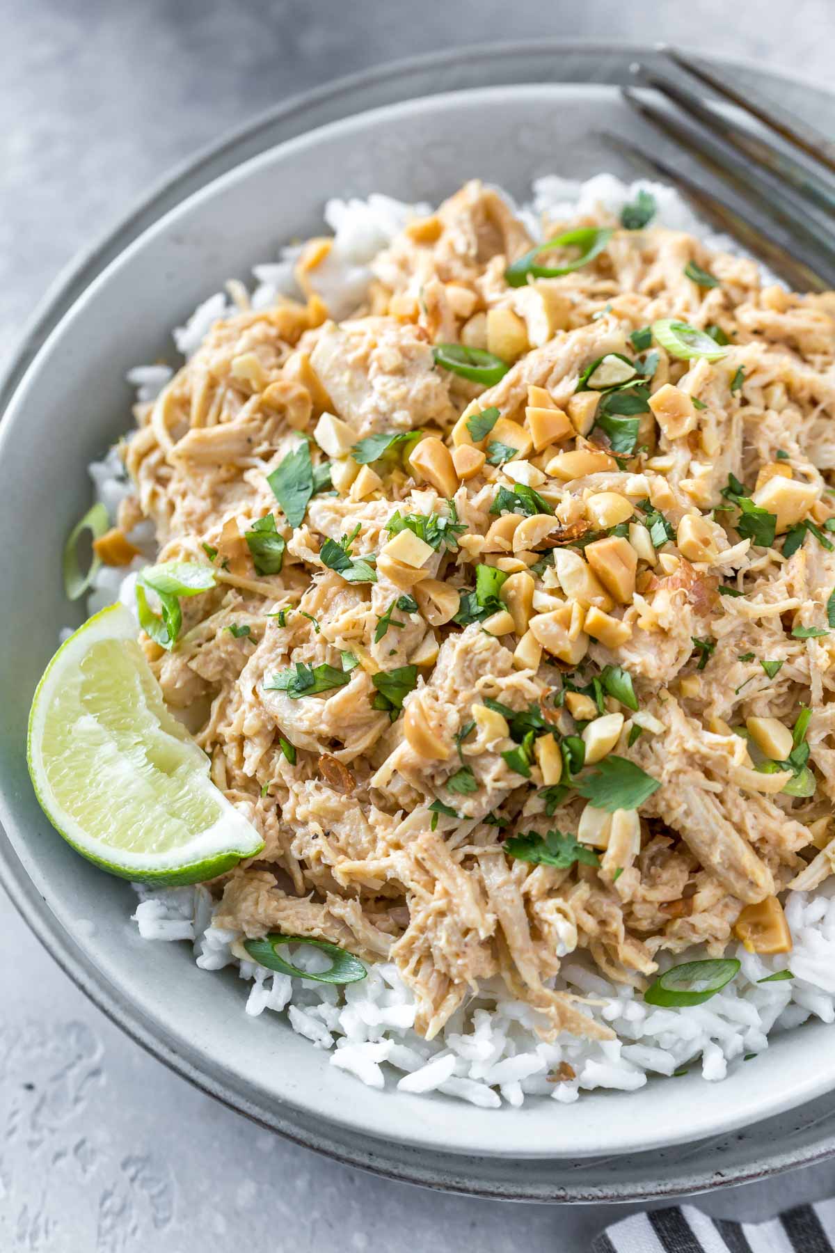closeup shot of shredded thai peanut chicken on a bed of coconut rice garnished with a lime wedge, chopped peanuts and cilantro, in a shallow gray bowl on a gray background next to a black and white striped napkin, two lime wedges and a bowl of coconut rice in the corner