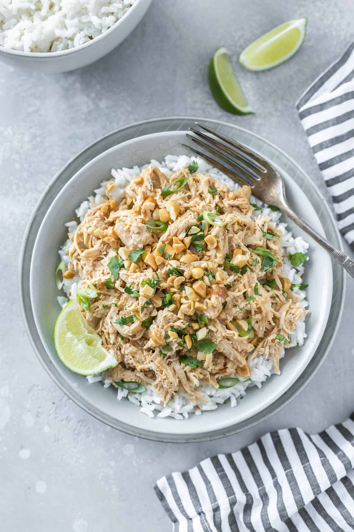overhead shot of shredded thai peanut chicken on a bed of coconut rice garnished with a lime wedge, chopped peanuts and cilantro, in a shallow gray bowl on a gray background next to a black and white striped napkin, two lime wedges and a bowl of coconut rice in the corner