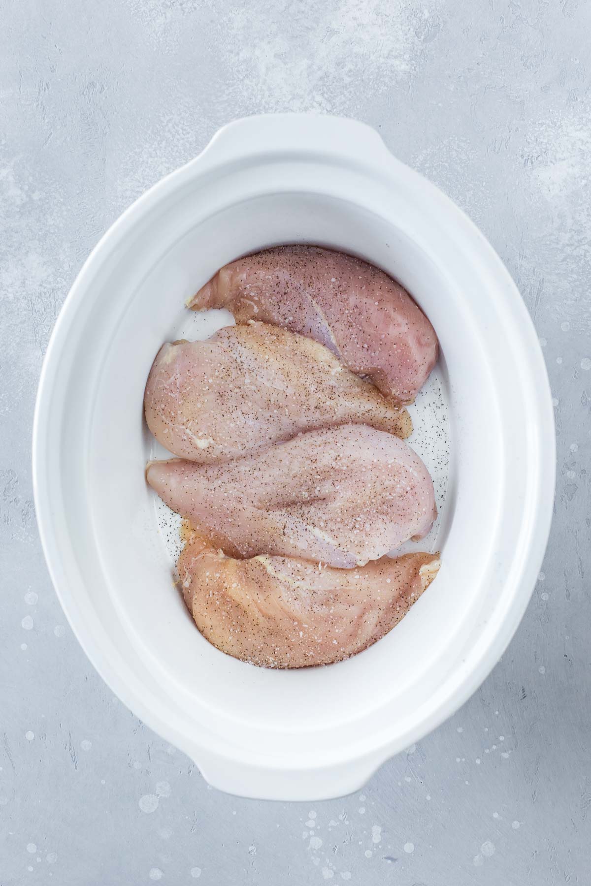 4 raw boneless, skinless chicken breast in a white slow cooker bowl on a gray surface, seasoned with salt & pepper