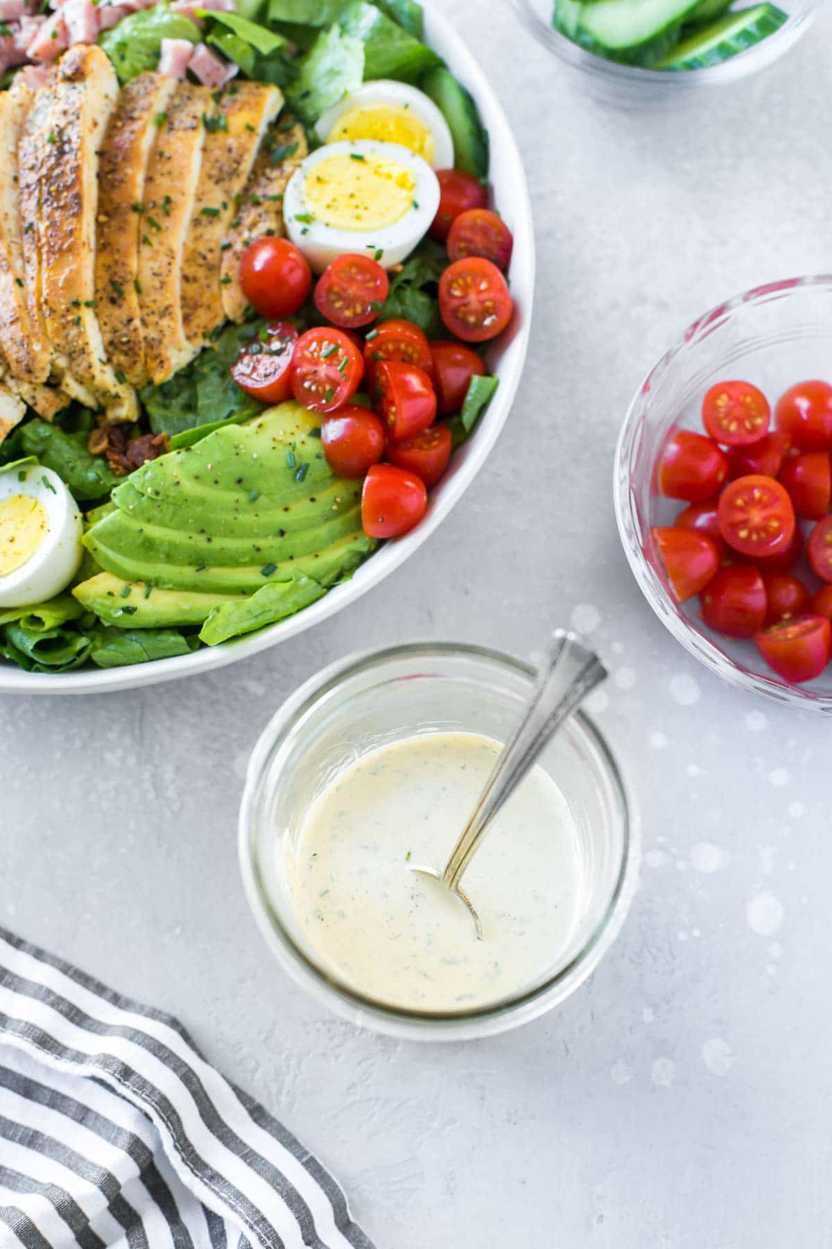 overhead view of a jar of dairy free ranch dressing with a spoon in it, a chicken cobb salad in a large bowl and some bowls with chopped veggies sitting behind it, a black and white striped napkin in the foreground