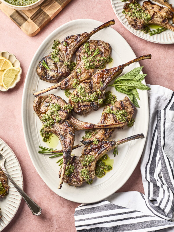 Grilled lamb chops, prepared with a flavorful lamb chop marinade, are arranged on an oval platter and topped with green herb sauce. Lemon slices, a striped napkin, and plates with more chops and sauce sit nearby on a pink surface.