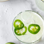 closeup of a spicy jalapeno margarita topped with three jalapeño slices on a white marble surface