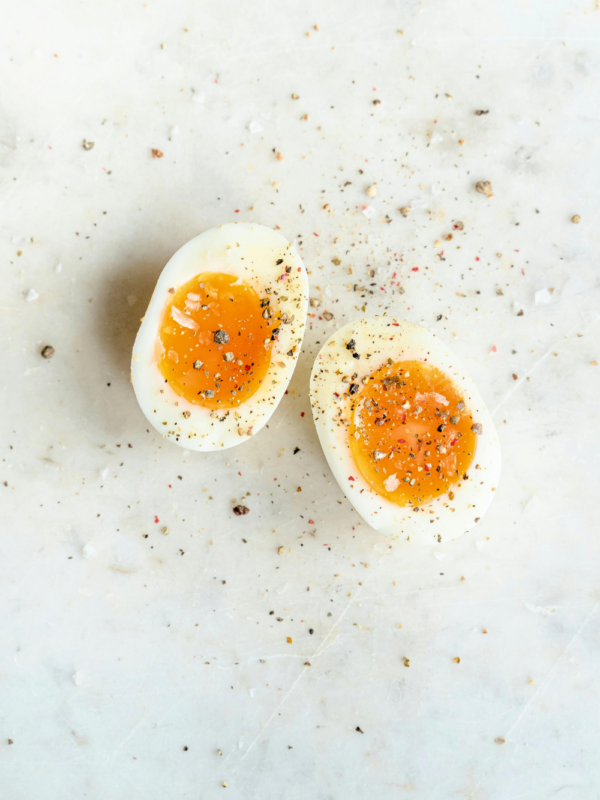 A soft-boiled egg cut in half, showing a bright orange, slightly runny yolk, sprinkled with salt, pepper, and seasoning on a light marble surface—reminding us that eggs are not dairy.