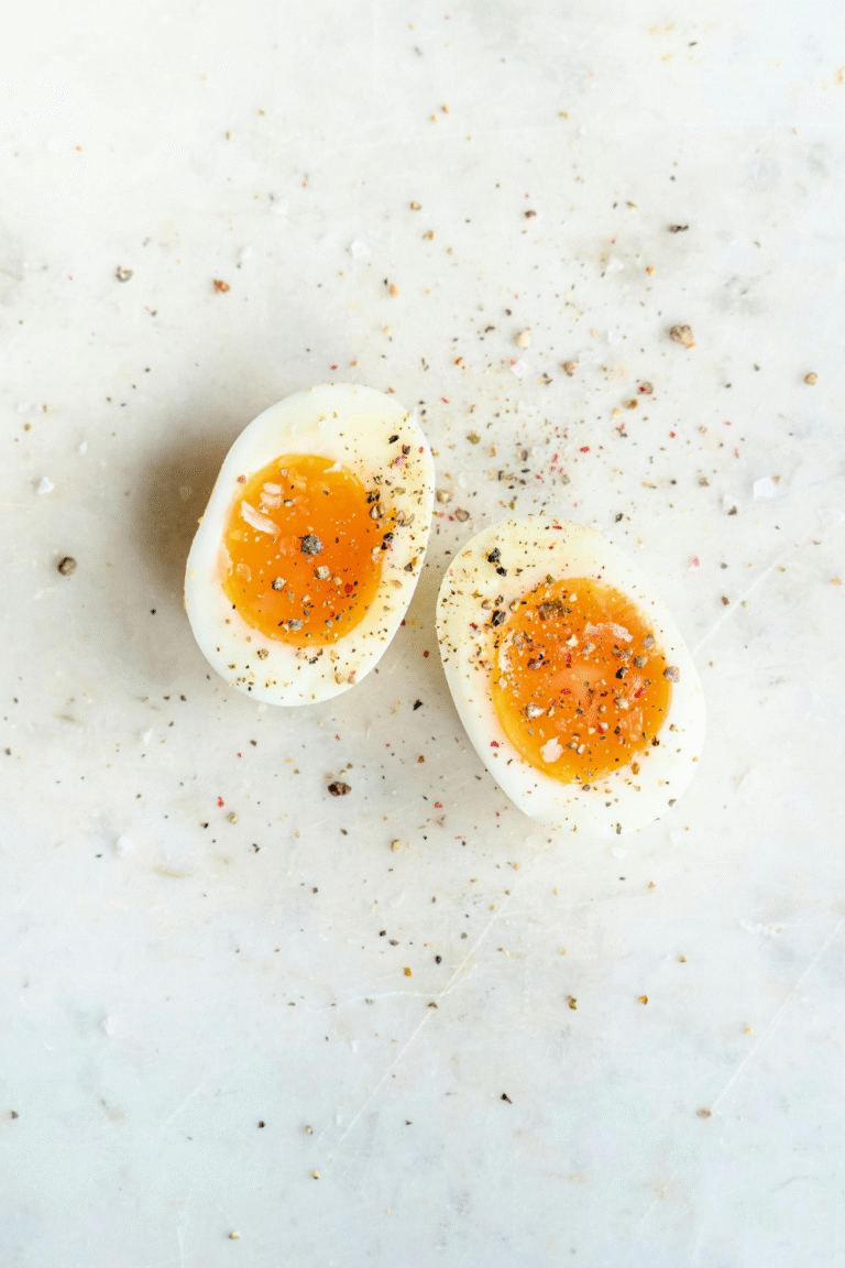 A soft-boiled egg cut in half, showing a bright orange, slightly runny yolk, sprinkled with salt, pepper, and seasoning on a light marble surface—reminding us that eggs are not dairy.