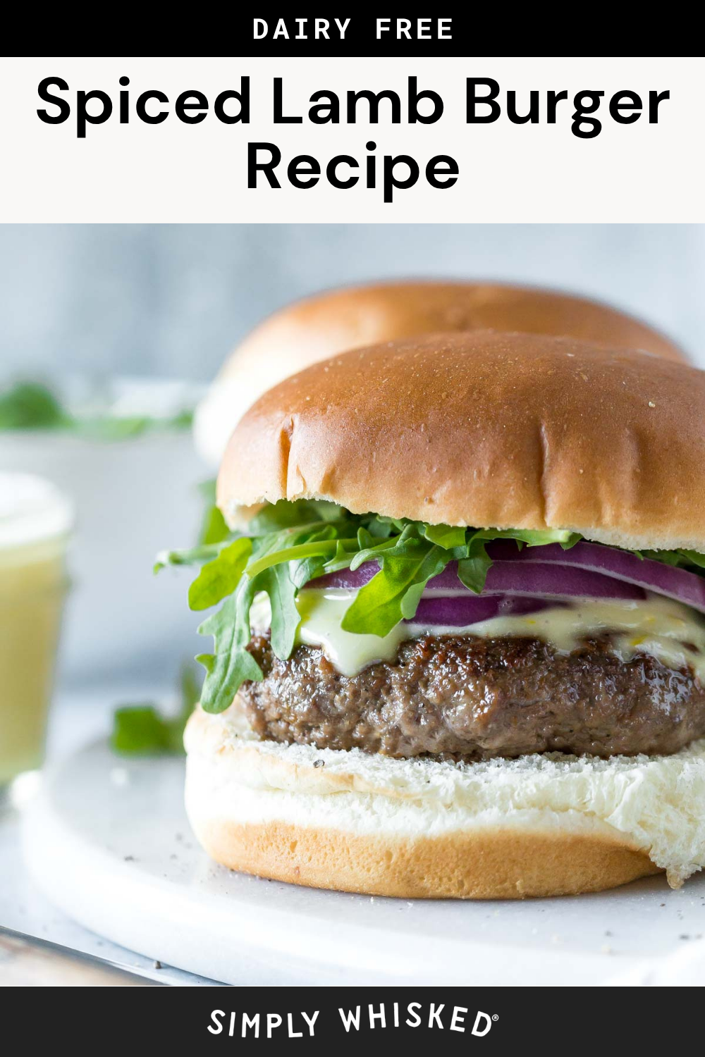 Close-up of a spiced lamb burger on a bun with lettuce, red onion, and sauce. The text above reads Dairy Free Spiced Lamb Burger Recipe. Another burger is blurred in the background, making this delicious lamb burger recipe look even more irresistible.