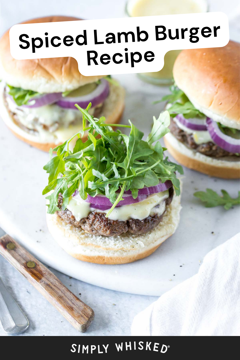 Two lamb burgers on buns topped with melted cheese, red onion, and arugula are on a white plate. Text at the top reads Spiced Lamb Burger Recipe, making this the perfect recipe for lamb burgers. A knife and fork are nearby.