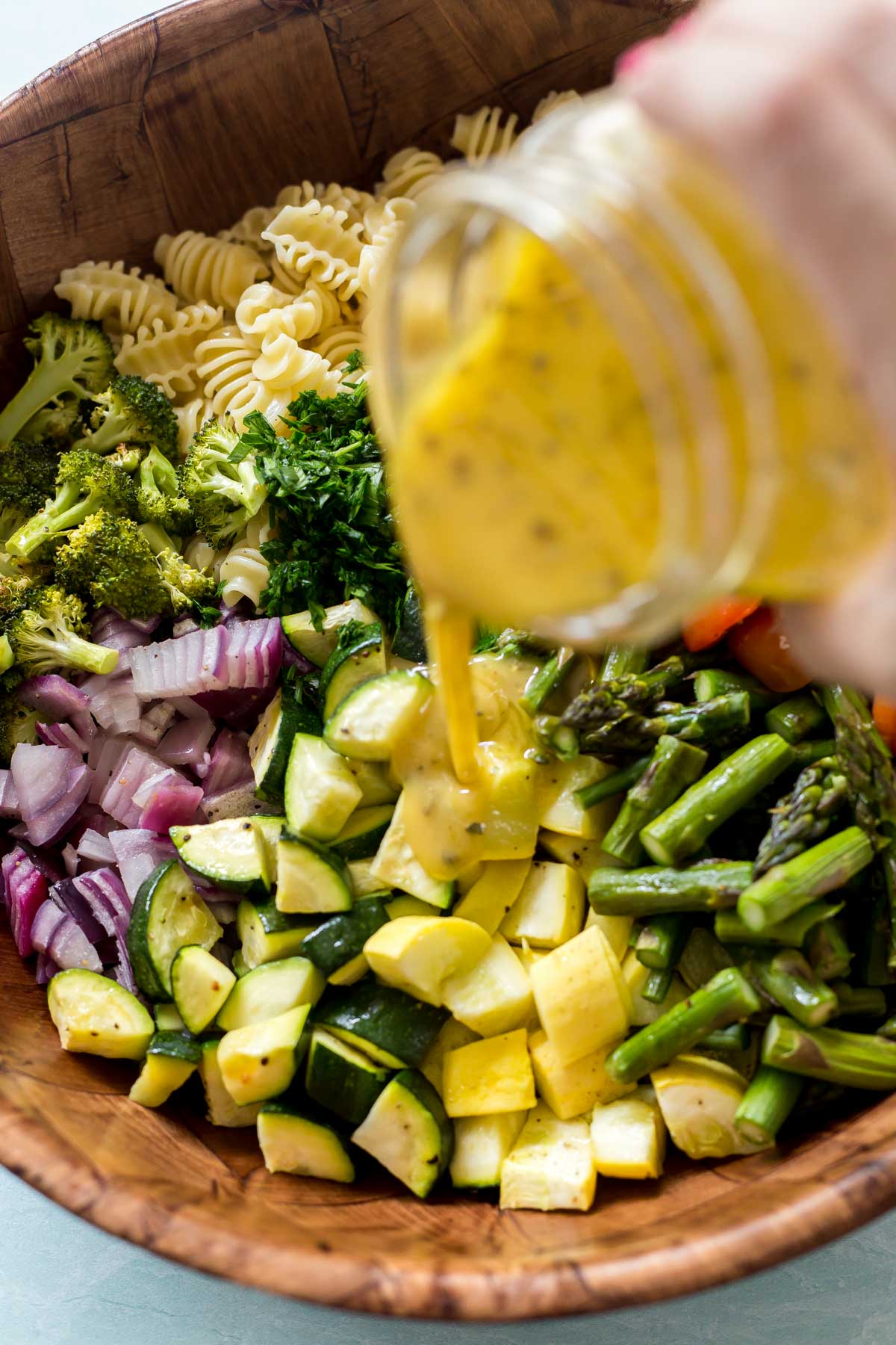 pouring homemade pasta salad dressing into a bowl with the ingredients for a roasted vegetable pasta salad recipe