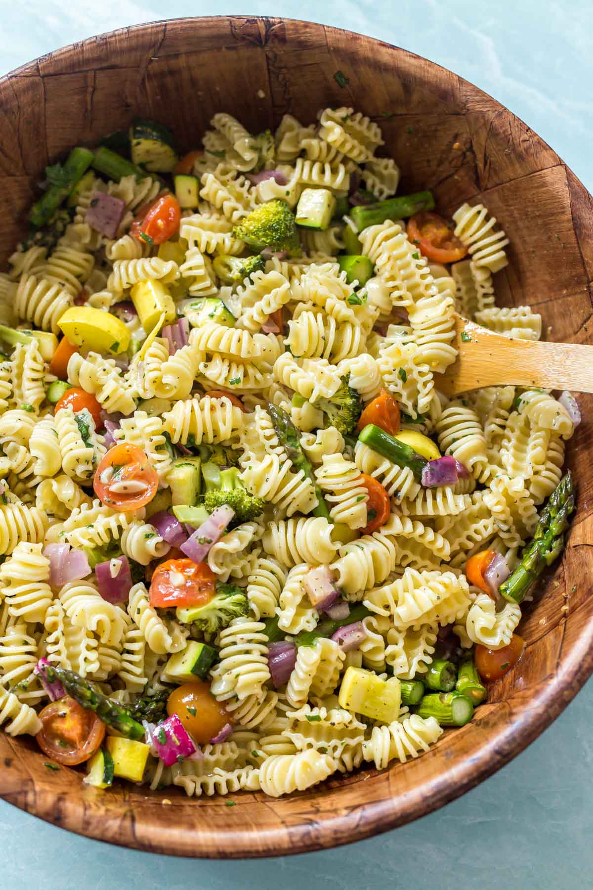 Roasted vegetables and cooked pasta being tossed with pasta salad dressing in a large bamboo bowl