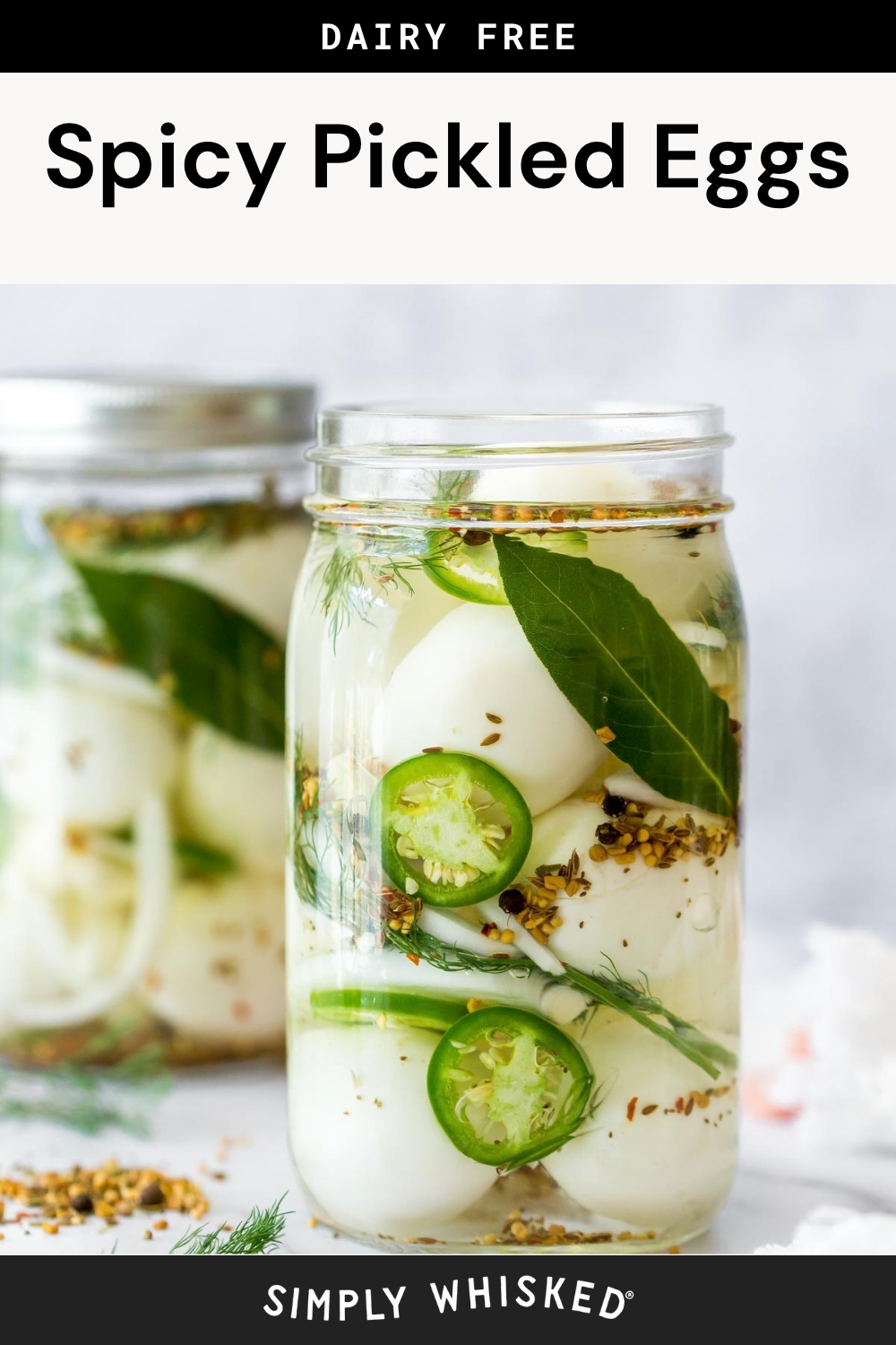 A jar filled with spicy pickled eggs, sliced jalapenos, bay leaves, herbs, and spices sits in focus, while another similar jar is blurred in the background. The label reads Spicy Pickled Eggs.