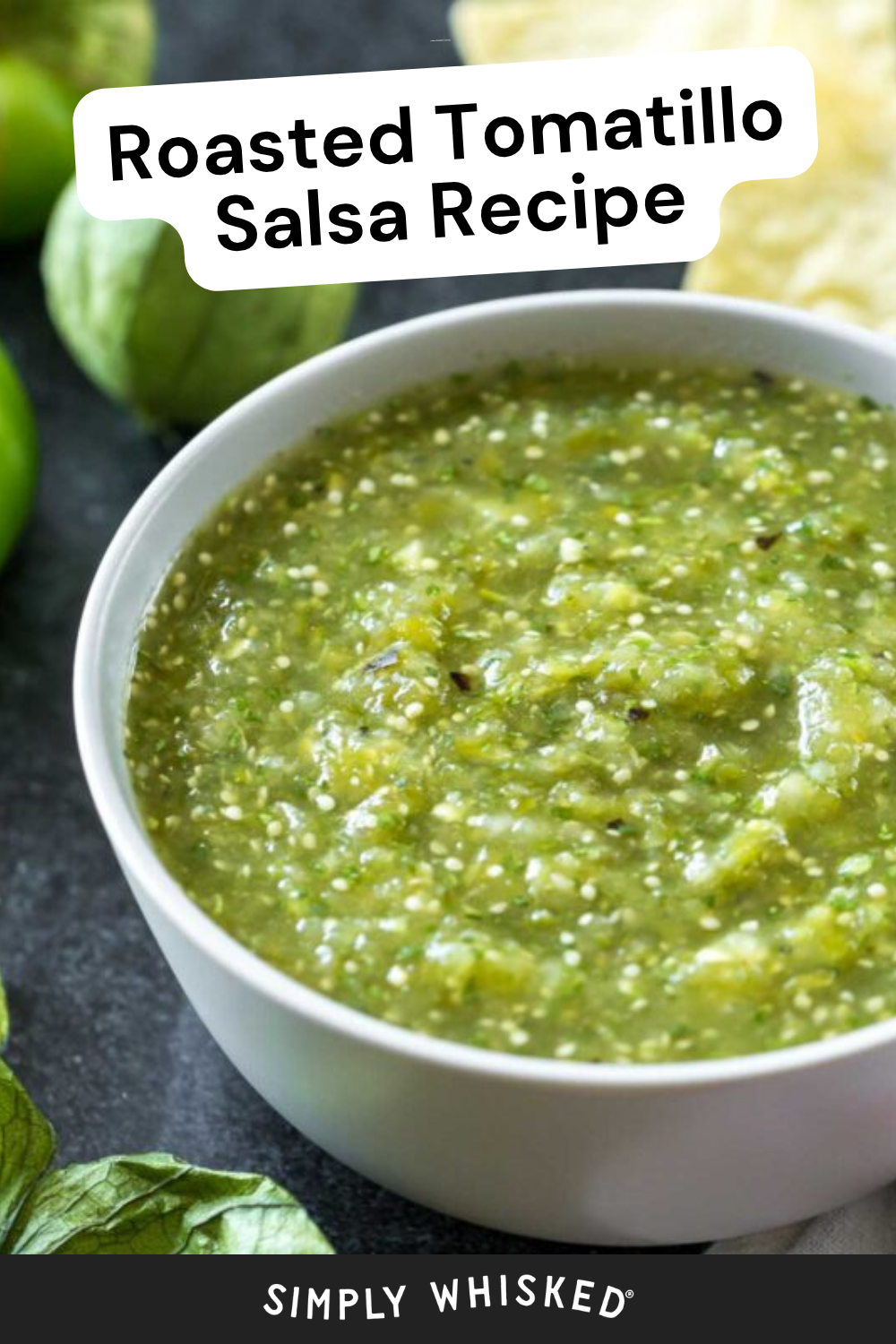 A bowl of vibrant roasted tomatillo salsa sits on a dark surface with fresh tomatillos and crispy tortilla chips nearby. Text reads Roasted Tomatillo Salsa Recipe and Simply Whisked at the bottom.