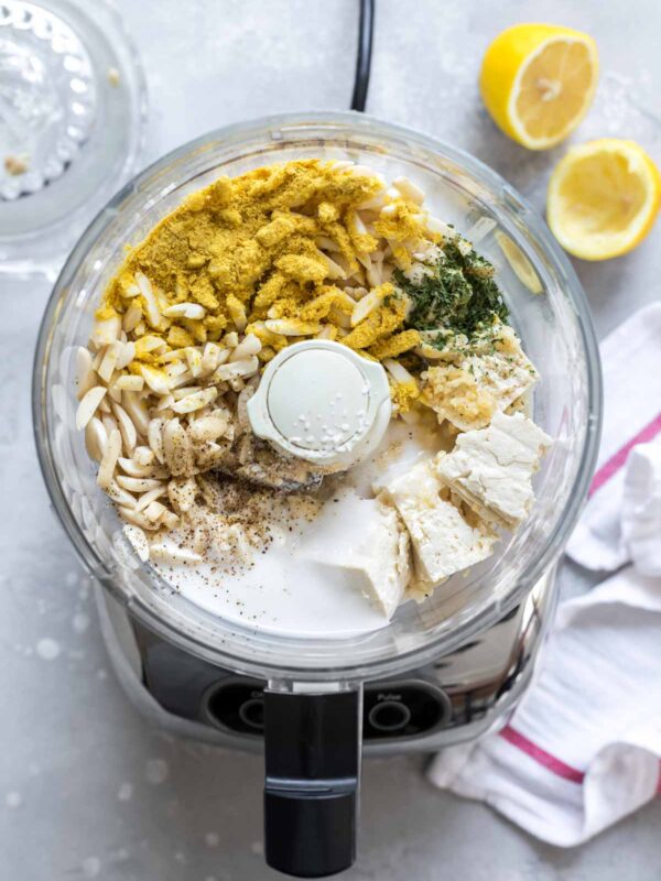 A food processor filled with slivered almonds, nutritional yeast, fresh herbs, black pepper, and chunks of vegan ricotta cheese, with halved lemons and a striped towel nearby on a light countertop.
