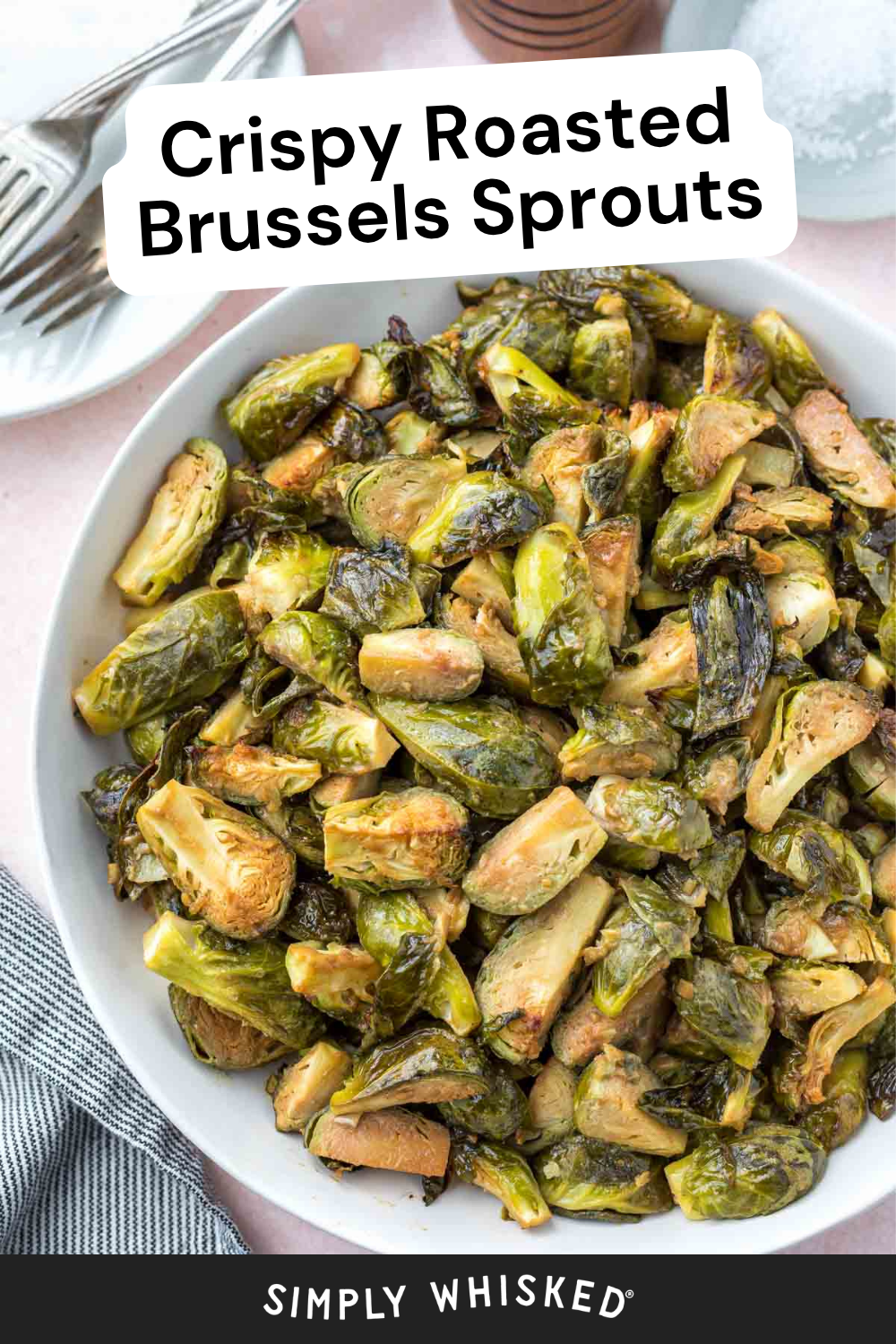 A bowl filled with perfectly roasted Brussels sprouts sits on a table with a striped napkin, a fork, knife, and a salt shaker nearby. Text reads Crispy Roasted Brussels Sprouts and Simply Whisked.