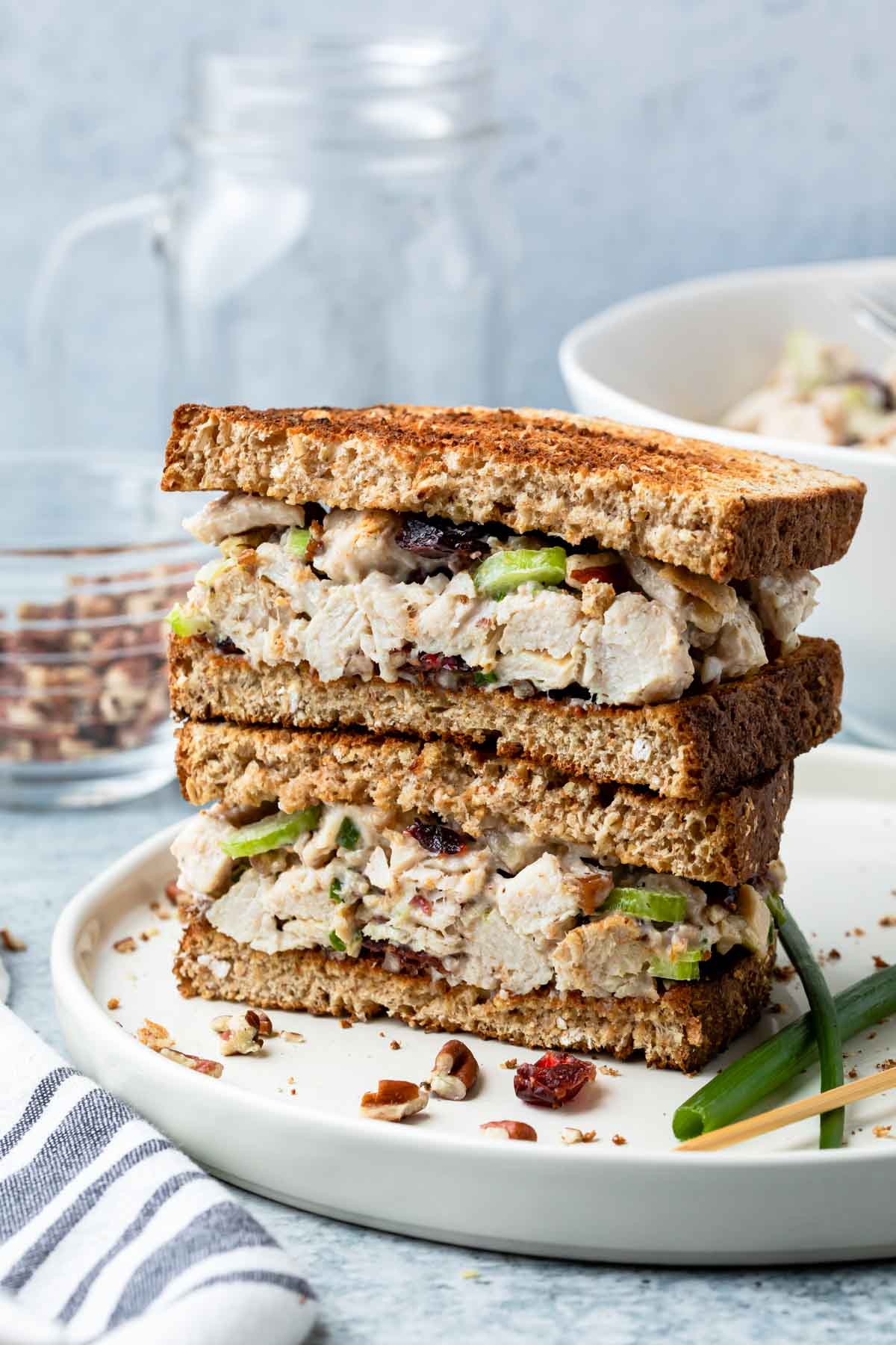Two halves of a turkey salad sandwich with greens and nuts are stacked on a white plate, with a bowl, glass, and jar blurred in the background. A striped napkin and green onion garnish are beside the plate.